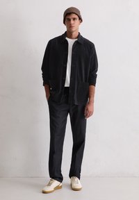 Donkergrijze corduroy overhemdjack met een kraag, knopen aan de voorkant en zakken, gecombineerd met zwarte broek en off-white sneakers. Bruine gebreide beanie.