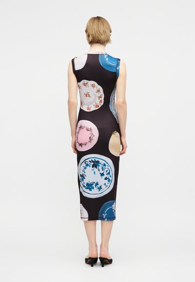 Γυναίκα που φοράει μαύρο midi φόρεμα χωρίς μανίκια με πολύχρωμα διακοσμητικά σχέδια, στέκεται με την πλάτη προς το μέρος μας, φορώντας μαύρες γόβες, σε λευκό φόντο.