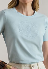 Frau trägt hellblaues T-Shirt mit erhabenem, kreisförmigem Ralph Lauren-Logo, kombiniert mit offwhitefarbenen Hosen und goldenen sowie Schildpatt-Armbändern.