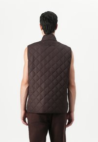 Καφέ, χωρίς μανίκια, επενδυμένο γιλέκο με διαμαντωτό σχέδιο, που διαθέτει ψηλό γιακά και διακριτικό λογότυπο στο πίσω μέρος. Λεία υφή, μινιμαλιστικός σχεδιασμός.