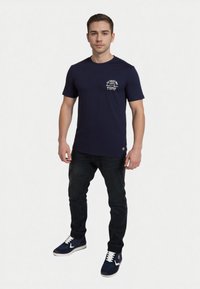 Camiseta azul marino de manga corta con un bolsillo en el frente y texto impreso en blanco. Combinada con jeans oscuros y zapatillas azules con acentos blancos.