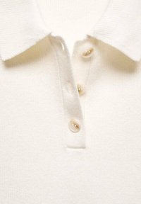 Col de chemise en maille blanche avec trois boutons beige clair sur une patte verticale, montrant la texture du tissu et les détails de couture.