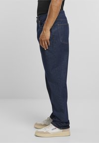 Mies, jolla on yllään tummansiniset farkut ja valkoiset lenkkarit, seisoo sivuttain yksinkertaista taustaa vasten.