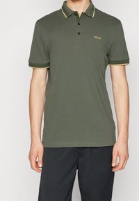 Man som bär en olivgrön kortärmad pikétröja med svart och gul kant på kragen och ärmarna, tillsammans med svarta byxor.