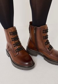 Brązowe skórzane botki za kostkę z sznurówkami i zamkiem błyskawicznym, teksturowana cholewka, okrągły nosek i masywna czarna gumowa podeszwa.