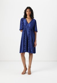Blue pleated dress with a deep V-neck and short puffed sleeves, featuring a cinched waist and flowing skirt, paired with brown strappy heels.