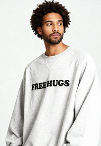 Sweat-shirt gris à coupe décontractée, arborant le texte noir « FREE HUGS » sur la poitrine. Confectionné en tissu doux et texturé avec des manches raglan.