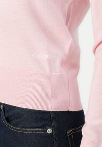 Maglione a maglia rosa chiaro con polsini e orlo a coste, caratterizzato da un piccolo logo bianco "Calvin Klein". Abbinato a jeans di denim scuri.