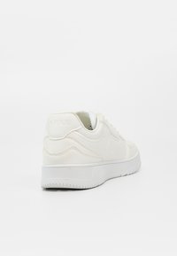 Baskets basses blanches avec une semelle épaisse et un motif subtil en forme de vague cousu, vues de l'arrière gauche sur un fond blanc uni.