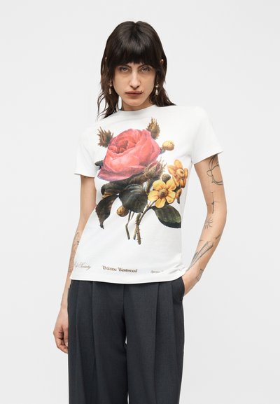 Γυναίκα με σκούρα μαλλιά που φοράει ένα λευκό T-shirt με μεγάλα ροζ και κίτρινα γραφικά λουλουδιών και σκούρο γκρι παντελόνι, στέκεται με το ένα χέρι στην τσέπη.