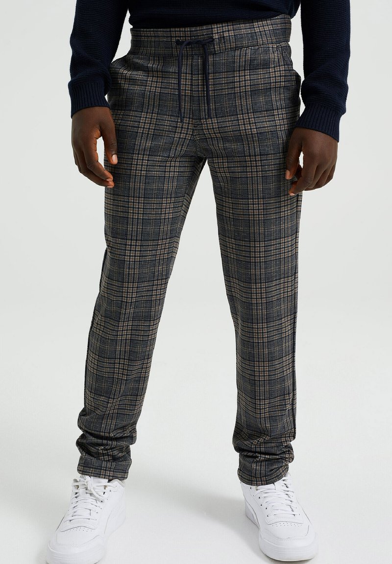 Homme portant un pantalon à carreaux gris et noir avec une taille élastique et un cordon, associé à des baskets blanches et un haut sombre à manches longues.