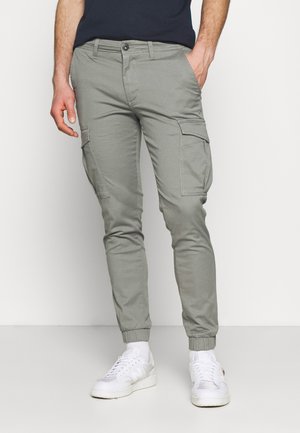 Mand iført lysegrå cargo joggebukser med elastiske manchetter og hvide sneakers, stående med den ene hånd i lommen.