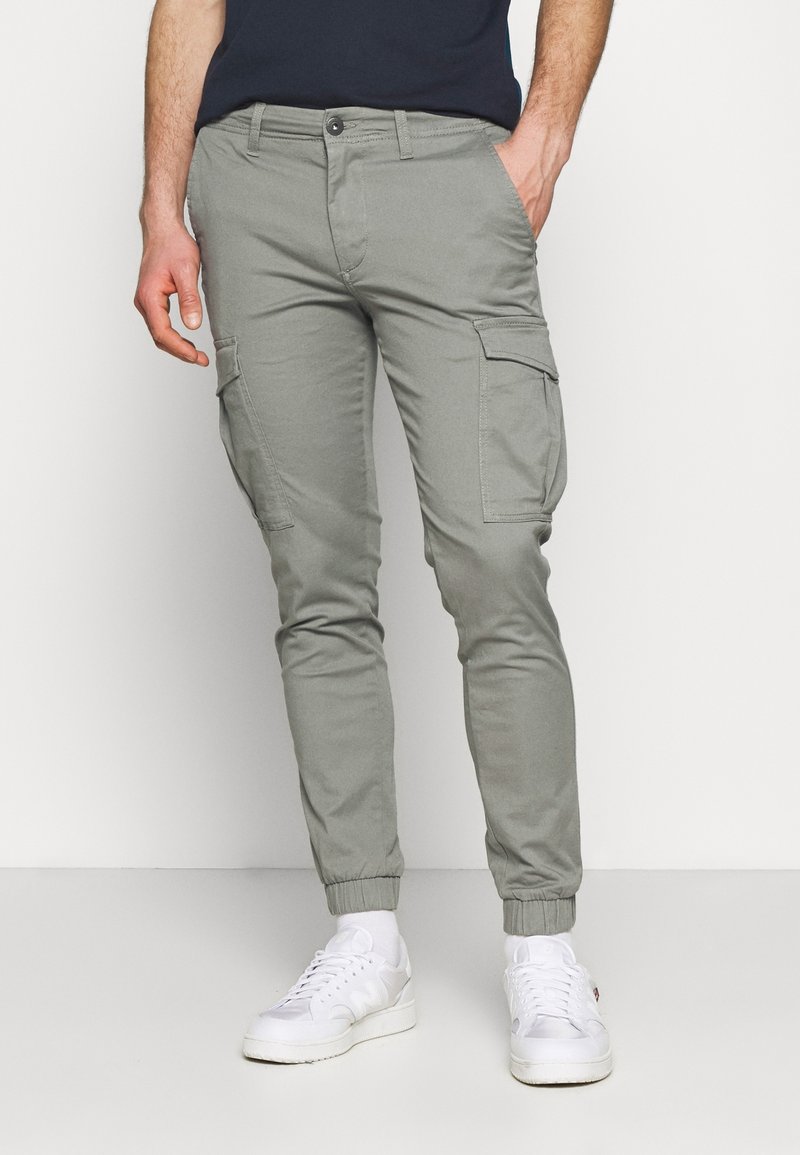 Homme portant un pantalon jogger cargo gris clair avec des poignets élastiques et des baskets blanches, debout avec une main dans la poche.