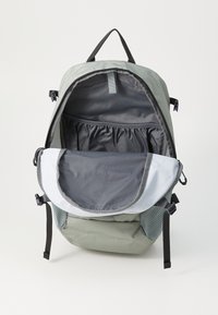 Zaino in tessuto verde con interni grigi. Presenta tasche laterali in rete, una parte superiore piatta, spallacci regolabili e un'apertura con patta per l'accesso.