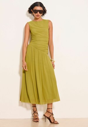 Woman wearing sleeveless olive green midi dress with ruched waist, dark sunglasses, gold hoop earrings, gold bracelet, and strappy brown high-heeled sandals.