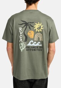 Olijfgroene katoenen t-shirt met een grafisch ontwerp dat een palmboom, zon, gezicht, regenboog, cactus en de tekst "BALANS VAN TEGENPOLES" bevat.