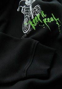 Sweat-shirt noir avec un graphique de squelette blanc faisant du vélo et du texte vert disant "Keep it Real" au-dessus de la Manche côtelée.