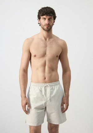 Uomo con capelli scuri e pelle chiara, senza maglietta, indossa pantaloncini chiari con coulisse, in piedi contro uno sfondo bianco semplice.