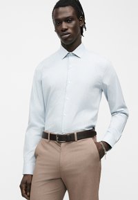 Άνδρας με ανοιχτό μπλε πουκάμισο με κουμπιά, ανοιχτό καφέ παντελόνι, μαύρη ζώνη και ασημένιο βραχιόλι, στέκεται με το ένα χέρι στην τσέπη σε λευκό φόντο.