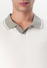 Cremfarbendes Poloshirt mit strukturiertem Stoff, grauem Kragen mit weißem Rand und kleinem gesticktem Logo auf der linken Seite.