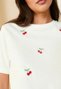 T-shirt blanc à manches courtes avec broderie de cerises rouges éparpillées et feuilles vertes. Tissu doux, encolure ronde, coupe décontractée.