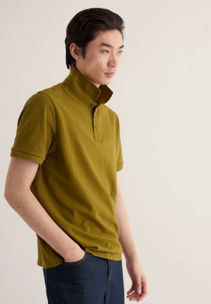 Giovane uomo con capelli neri che indossa una polo verde oliva con colletto alzato e jeans blu scuro, in piedi con una mano in tasca.