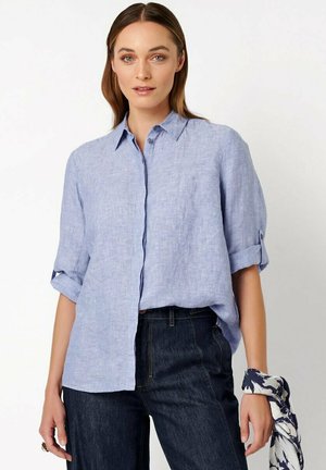 Donna che indossa una camicia di lino azzurro chiaro con bottoni e maniche arrotolate e jeans blu scuro a vita alta, tiene un foulard con motivo al polso.