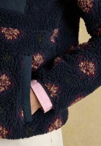 Fleece pullover in marineblauw met bloemenpatronen in tinten van bordeauxrood, roze en groen, voorzien van een donkerblauwe zak en een roze manchet.