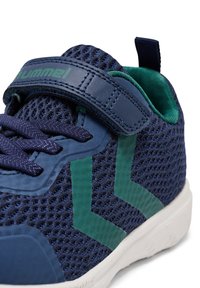 Blå nätidrottssko med gröna accenter, med en kardborreband och texturerad yta. Vit gummisula och intrikat snördetaljering.
