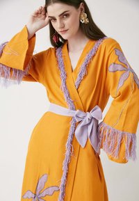 Túnica naranja con ribete de flecos lavanda y cinturón. Presenta patrones florales bordados en la parte delantera y las mangas. Tejido ligero.