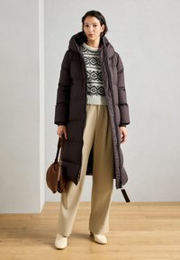 Manteau long matelassé marron avec une capuche, fermeture éclair, associé à un pull à motifs, un pantalon beige à jambes larges et des bottines crème.