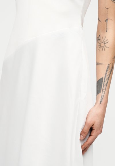 Κοντινό πλάνο ενός ατόμου που φοράει λευκό φόρεμα με εμφανή τατουάζ στο αντιβράχιο και το χέρι που ακουμπάει στο πλάι.