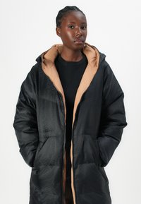 Black puffer coat with a tan lining, zip closure, two front pockets, and textured pattern on the exterior. Features a hooded design.