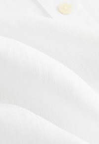 Tissu blanc à la texture lisse et légère, présentant des motifs tissés subtils et un seul bouton jaune partiellement visible.