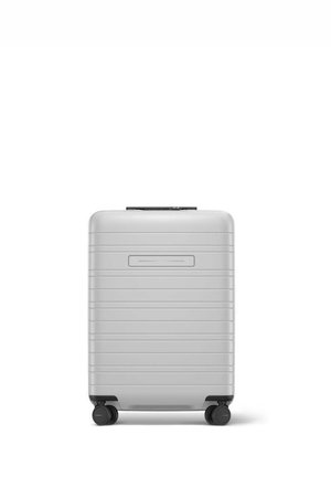 Valise rigide grise avec des rainures horizontales, poignée rétractable et quatre roues pour la mobilité. Texture lisse et design compact.