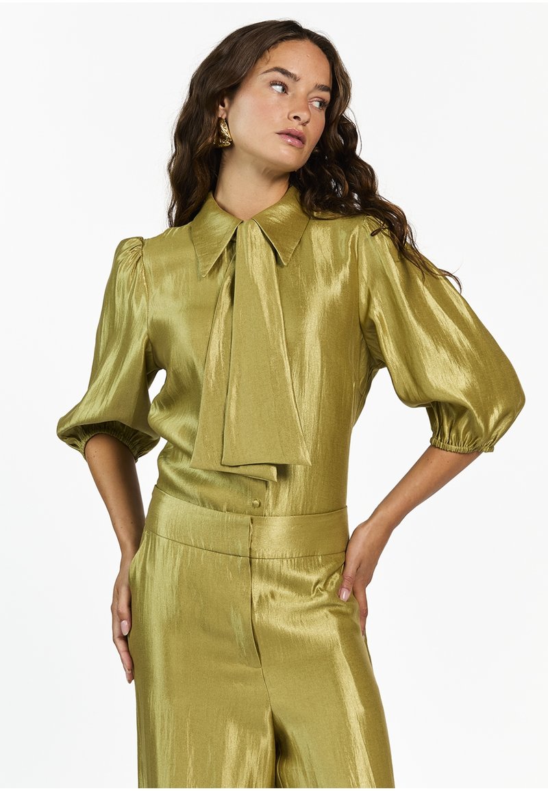 Olijfgroene, glanzende satijnen blouse met een grote kraag en strikdetail. Heeft puffed sleeves en een getailleerde taille. Zachte textuur.