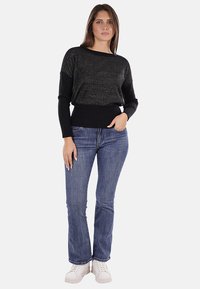 Maglione lavorato a maglia nero con un design a trama, orlo a coste e maniche nere, indossato con jeans a zampa d'elefante blu e sneakers bianche.