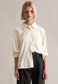 Kremfarget, langermet skjorte med knappestolpe og krage, rullet opp ermer, i kombinasjon med mørkegrå bukser. Har en glatt, blank tekstur.