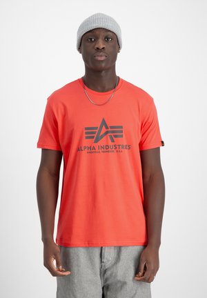 Crvena pamučna majica s crnim "Alpha Industries" logom i dizajnom; kratki rukavi; okrugli izrez; uparen s sivim hlačama i sivom kapom.