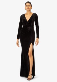 Langes schwarzes Samtkleid mit tiefem V-Ausschnitt, langen Ärmeln und einem seitlichen Schlitz. Hat ein figurbetontes Design mit subtilen Drapierungen an der Taille.