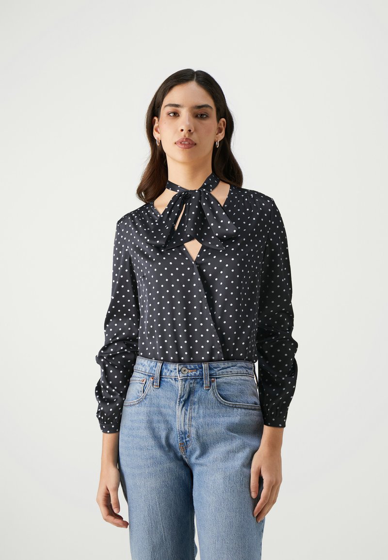 Blusa nera con pois, colletto a fiocco, con un dettaglio a chiave di volta e maniche lunghe. Abbinata a jeans azzurri a vita alta.