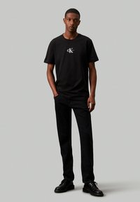 Camiseta de algodón negra con un pequeño logo blanco en el pecho, combinada con jeans negros y zapatos negros, sobre un fondo neutro.