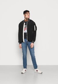 Jaqueta bomber preta com fecho branco, t-shirt gráfica, jeans azul claro e ténis brancos com detalhes em bordeaux. Modelo está contra um fundo simples.