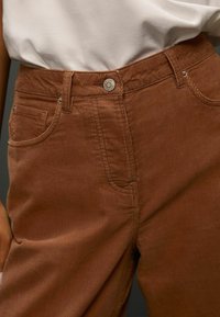 Braune Cordhosen mit vertikalen Rippungen, einer Vordertasche und einem Metallknopfverschluss. Weicher Stoff mit verstärkten Nahtdetails.