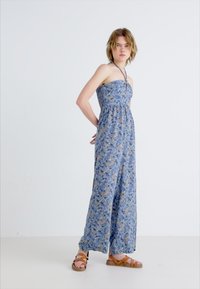 Modra cvetna maksi obleka brez naramnic, lahka tkanina in ohlapen kroj. Rjavi sandali s trakovi dopolnjujejo obleko.