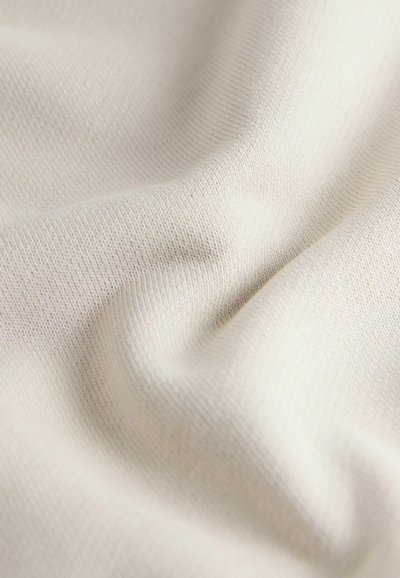 Gros plan sur un tissu doux de couleur crème avec une texture fine en maille côtelée et de légers plis créant des ombres subtiles.