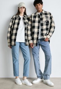 Juodi ir kremiškai dryžuoti flaneliniai marškiniai, džinsiniai džinsai, balti sportiniai bateliai ir kepurė. Tekstūruota medžiaga su kišenėmis ir matomais mygtukų detalėmis.