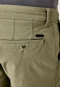 Olivgrüne Hose aus strukturiertem Stoff, mit einer Gesäßtasche und einem Knopfdetail sowie kontrastierenden schwarzen Akzentnähten.