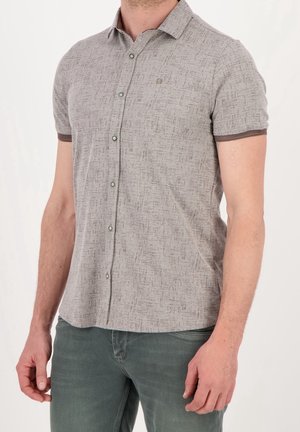 Mężczyzna ubrany w koszulę na guziki z krótkim rękawem w jasnoszary wzór z ciemnym obramowaniem na rękawach oraz zielone spodnie na jednolitym tle.