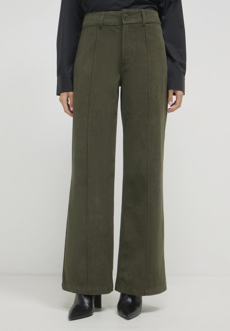 Pantalons en velours côtelé à jambes larges de couleur vert foncé, avec des côtes verticales, une fermeture à bouton et une taille haute. Associés à des escarpins noirs à bout pointu.